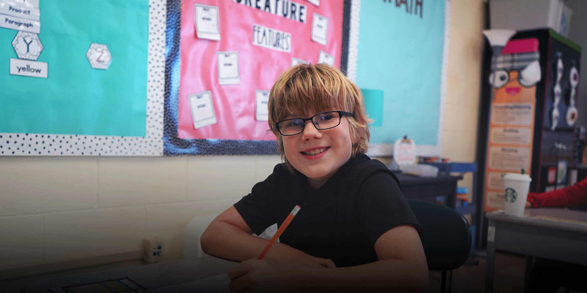 Smiling student in glasses writing at a classroom desk.