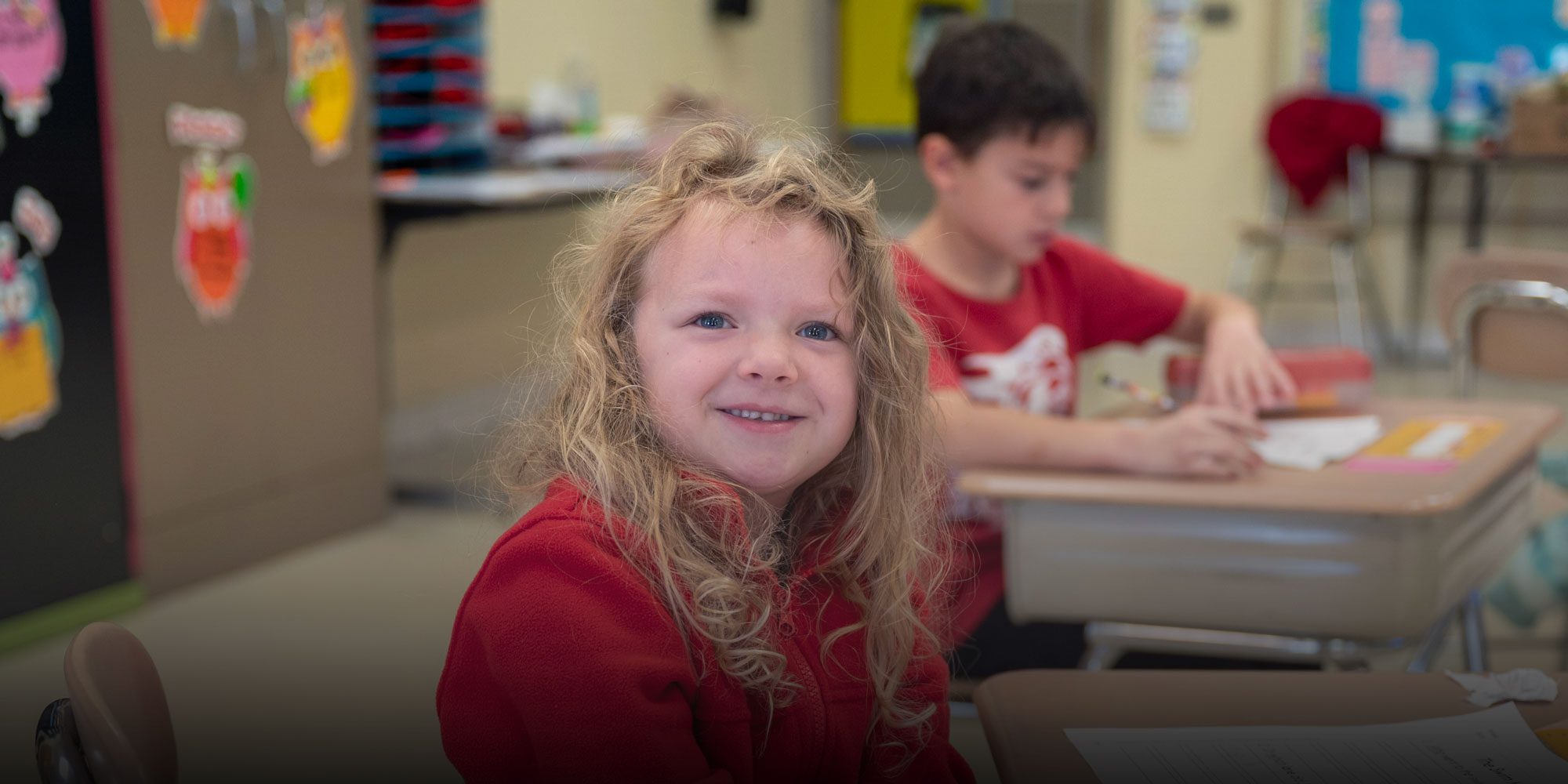Smiling student with curly blonde hair at a classroom desk.
